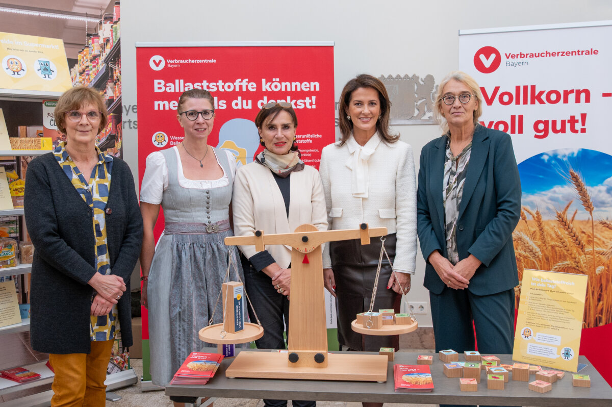 Ministerin Kaniber gemeinsam mit vier Frauen vor Informationsplakaten der Verbraucherzentrale Bayern zum Thema Ballaststoffe und Vollkorn, vor ihnen befindet sich ein Tisch mit einer Holzwaage und bunten Informationsmaterialien.