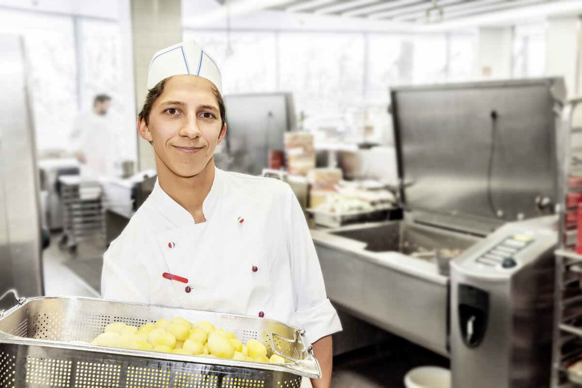 Person in Küchenkleidung hält Metallbehältnis mit Kartoffeln in einer Großküche