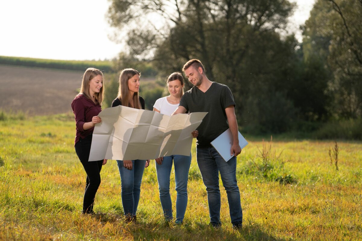 Drei junge Frauen und ein junger Mann stehen auf einer Wiese und betrachten einen Flurstückplan.