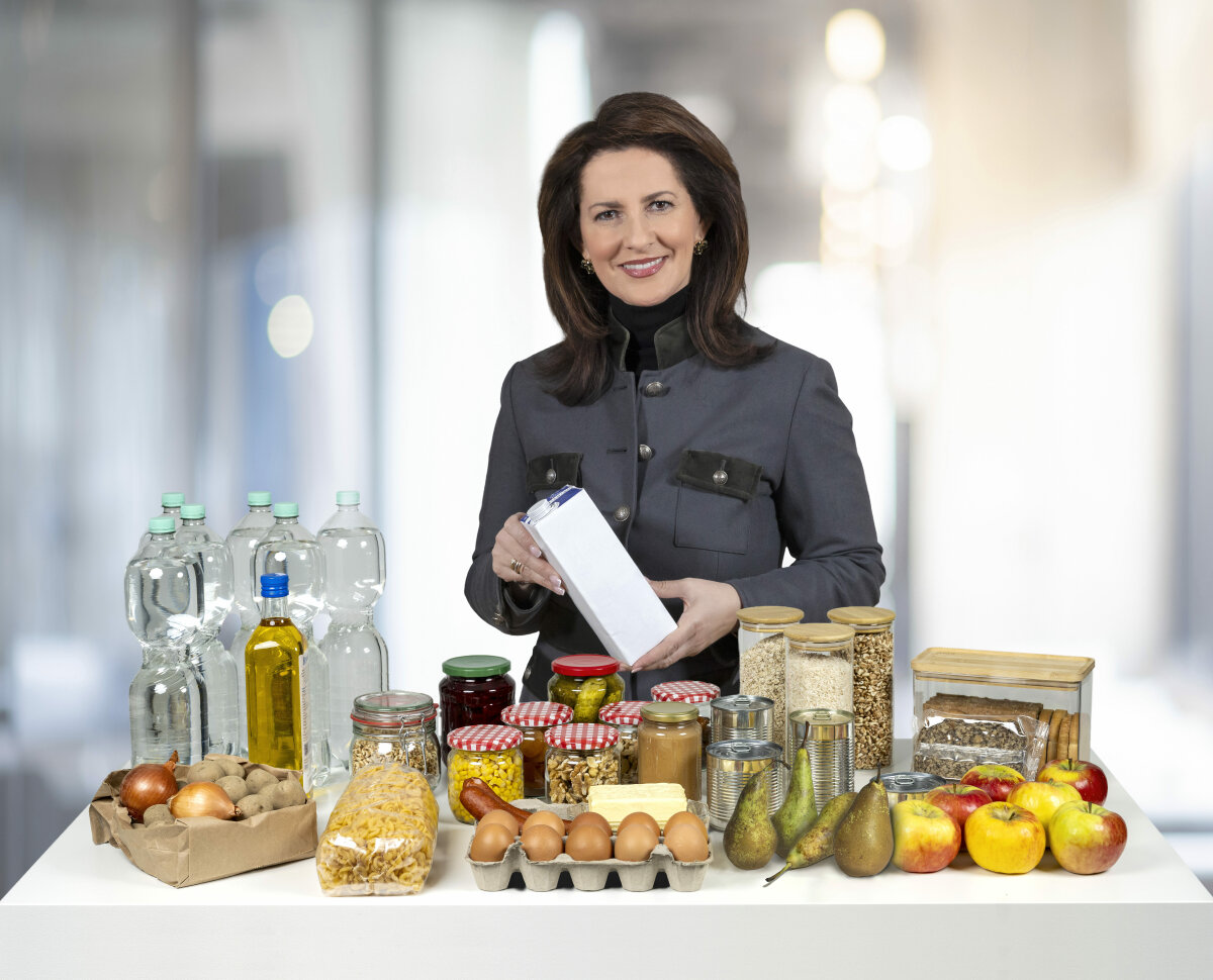 Staatsministerin Kaniber steht lächelnd hinter einem Tisch, auf dem diverse Lebensmittel stehen, z.B. Eier, Äpfel. Sie hält einen Milchkarton in der Hand.