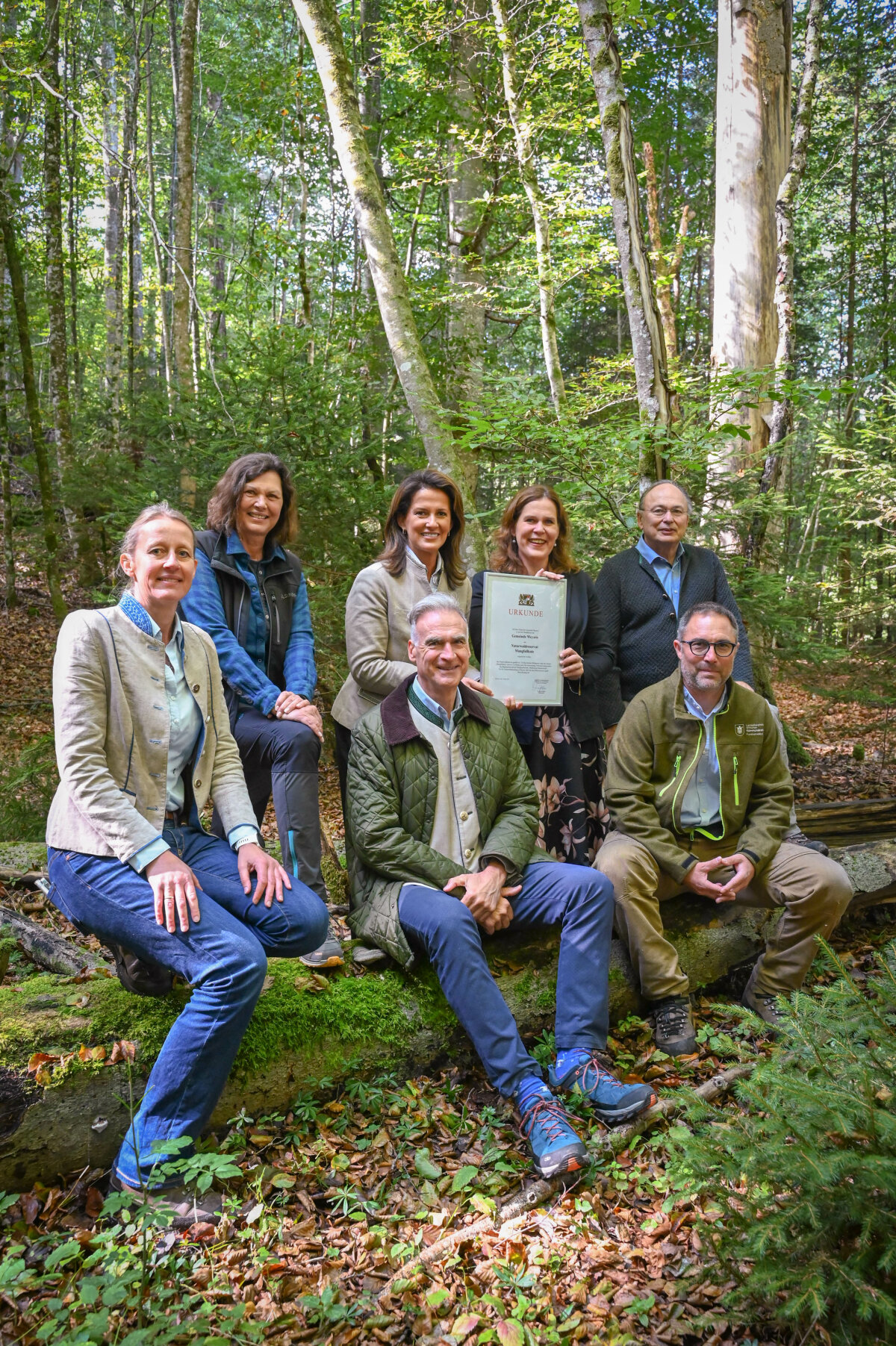 Staatsministerin Kaniber mit anderen Beteiligten in einem Waldstück. Einige der Personen sitzen auf einem Baumstamm, andere Personen stehen dahinter. 