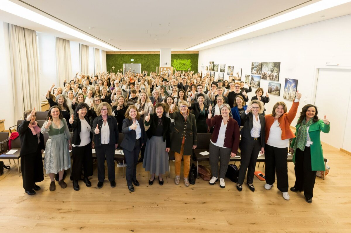 Die Absolventinnen und Absolventen stehen im Veranstaltungssaal des Ministeriums und lachen in die Kamera. Einige halten ihre Arme nach oben und zeigen den Daumen hoch.