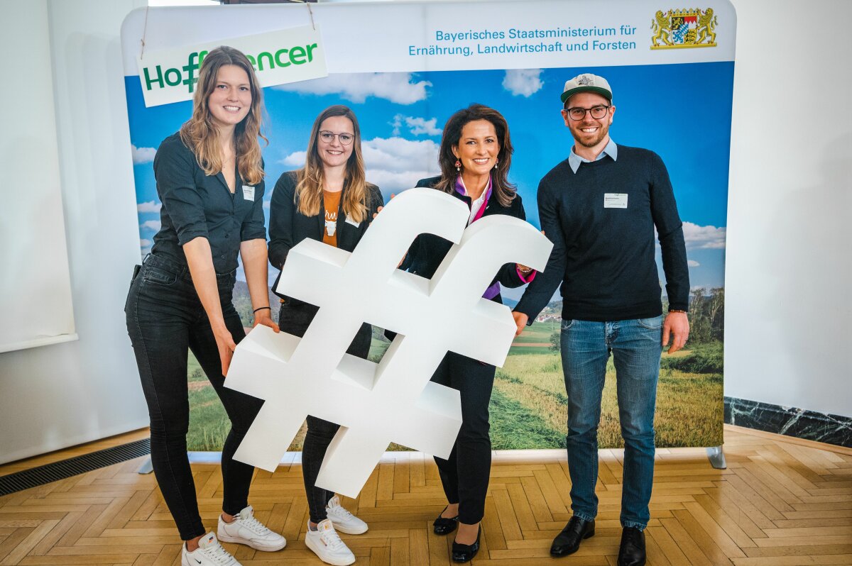 Ministerin Kaniber hält das Logo des Hoffluencer-Treffens in de Hand. Neben ihr stehen drei junge Teilnehmer der Veranstaltung