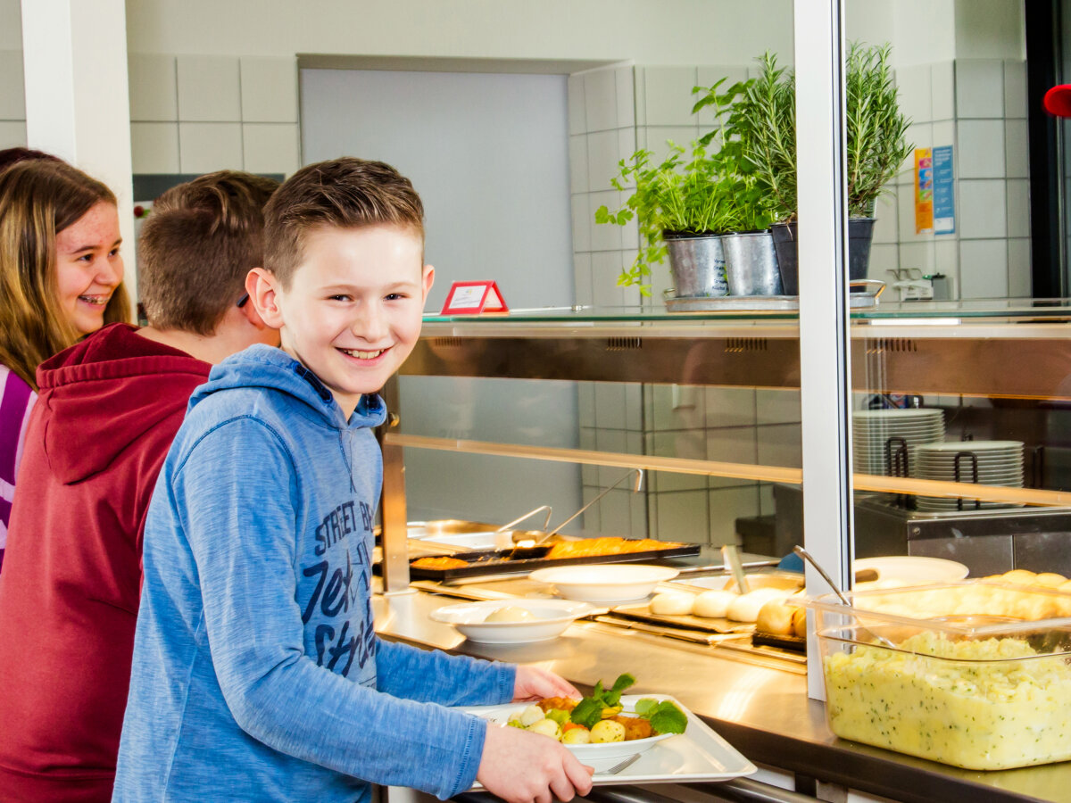 Mehrere Kinder stehen fröhlich an der Essensausgabe einer Mensa und erhalten ein warmes Essen auf Tabletts.