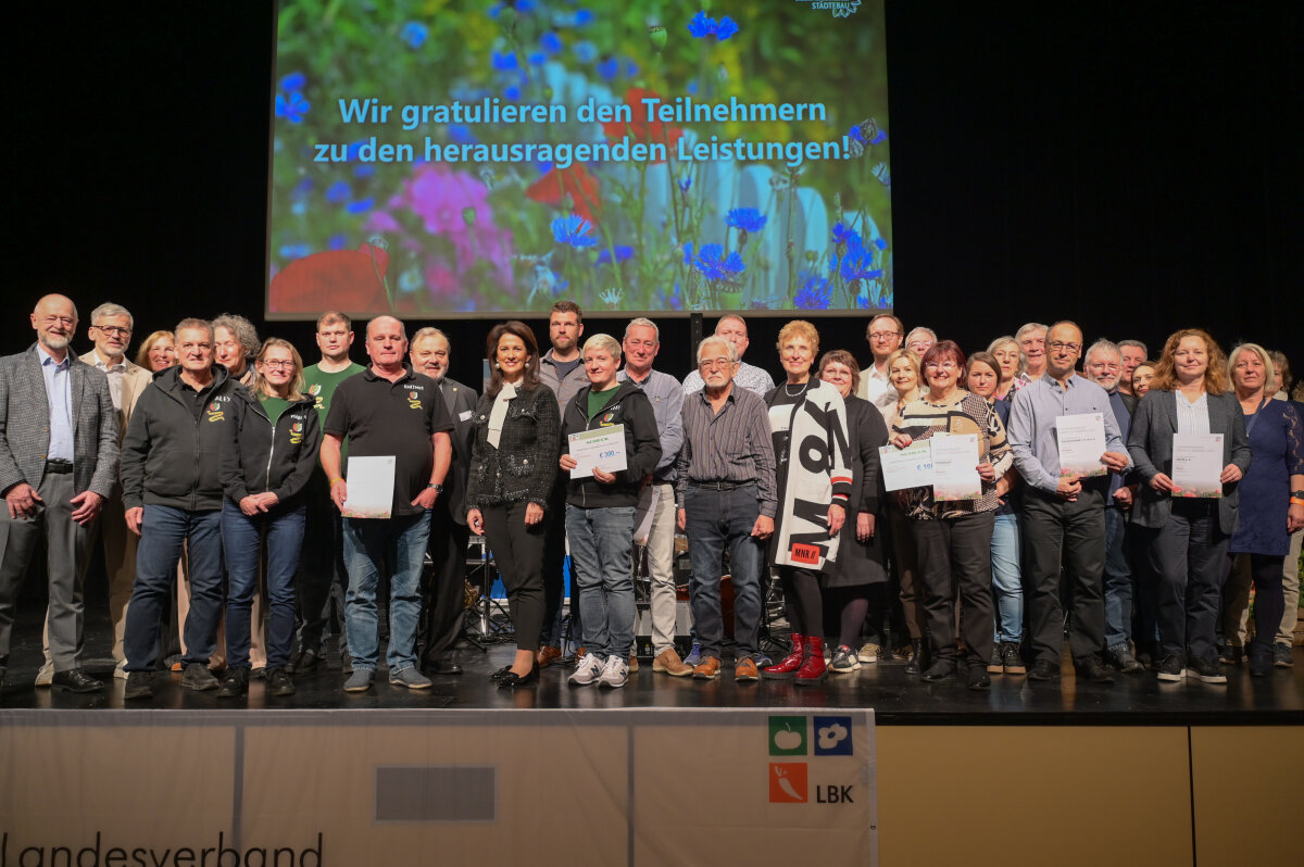 Die Wettbewerbsteilnehmer stehen lächelnd mit der Ministerin auf einer Bühne und halten Urkunden, während hinter ihnen eine Leinwand mit einem Glückwunschtext eingeblendet ist.