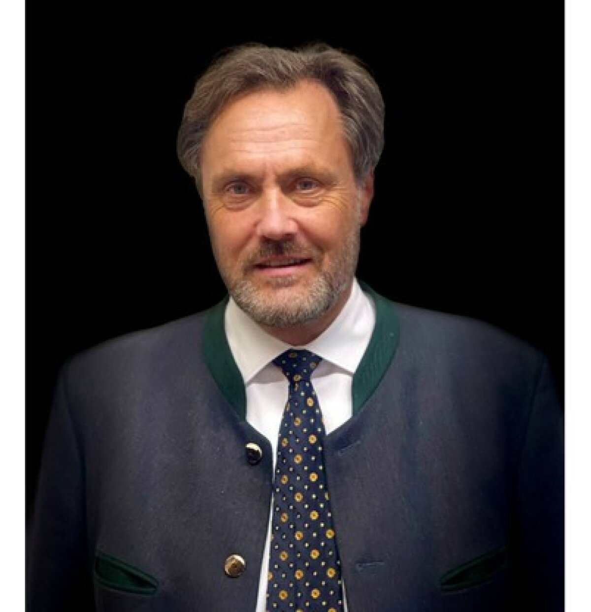 Dr. Neser lächelt in die Kamera vor einem schwarzen Hintergrund . Er trägt ein traditionelles bayerisches Sakko.