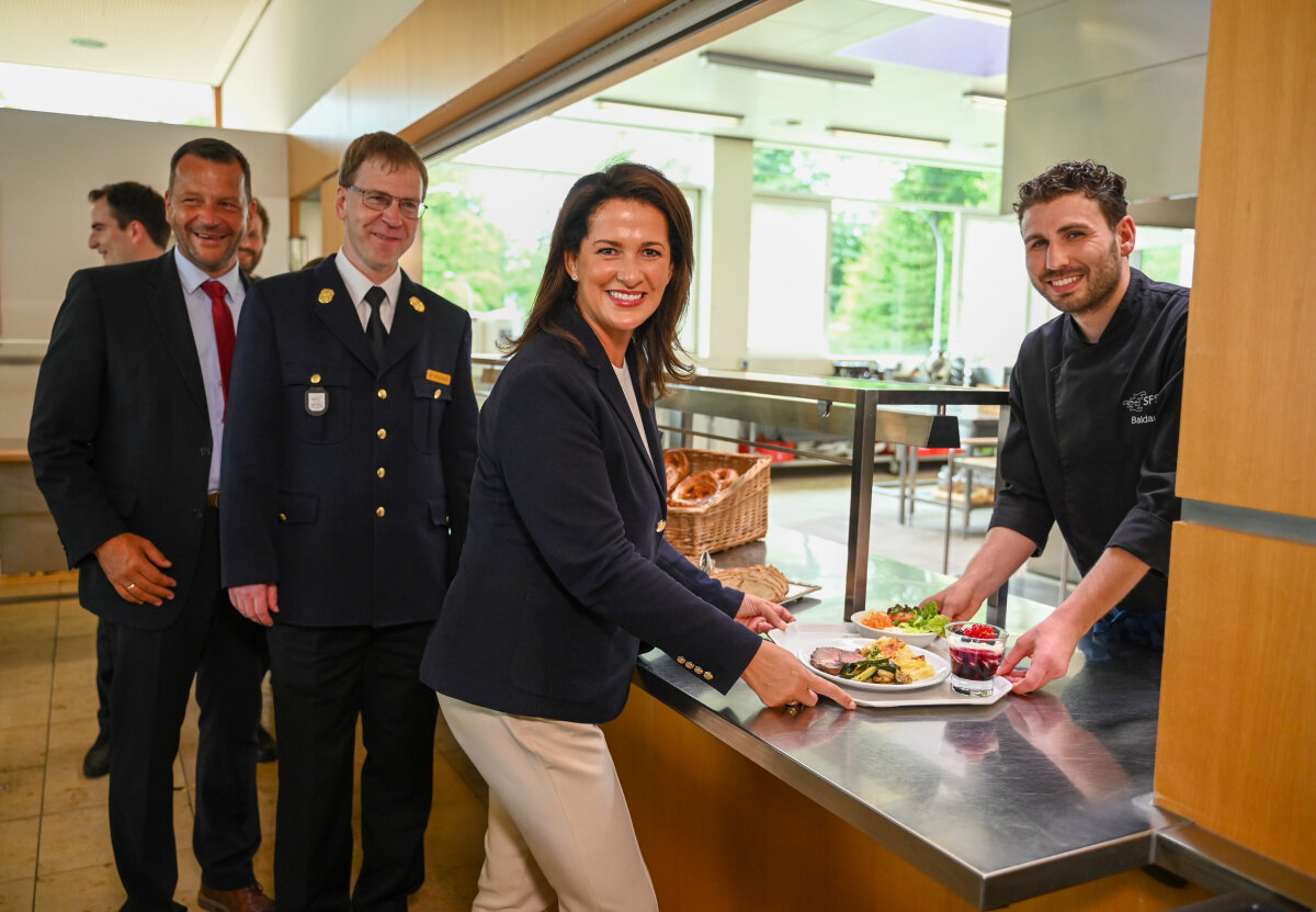 Auf dem Bild ist Ministerin Kaniber zu sehen, wie sie in einer Kantine ein Gericht von einem Koch entgegennimmt, während im Hintergrund weitere Personen, darunter auch ein Mann in Uniform, anstehen.
