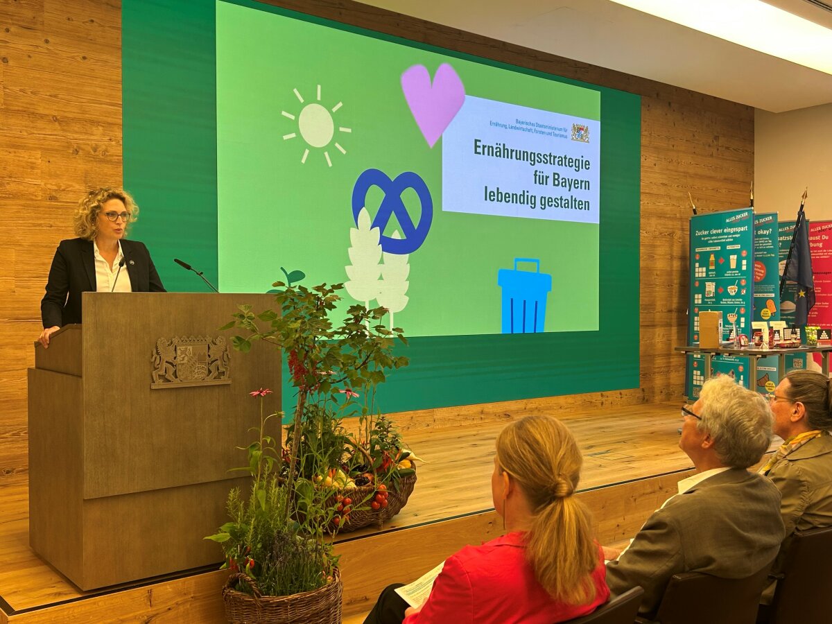 Auf dem Bild spricht Alexandra Schenk an einem Rednerpult im Veranstaltungssaal des Bayerischen Staatsministeriums. Hinter ihr zeigt eine große Leinwand den Titel „Ernährungsstrategie für Bayern lebendig gestalten“. Vor dem Pult stehen Körbe mit frischem Gemüse und Kräutern. Im Publikum sitzen mehrere aufmerksam zuhörende Personen.