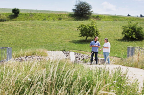 Ein Mann und eine Frau stehen auf einem Damm inmitten von Wiesen.