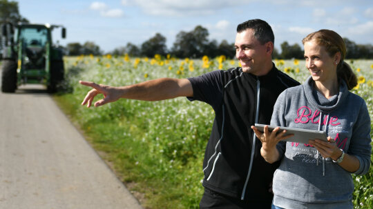 2 Personen stehen neben Feld: der Mann zeigt mit dem Finger nach links, die Frau hält ein Tablet; dahinter steht ein Traktor