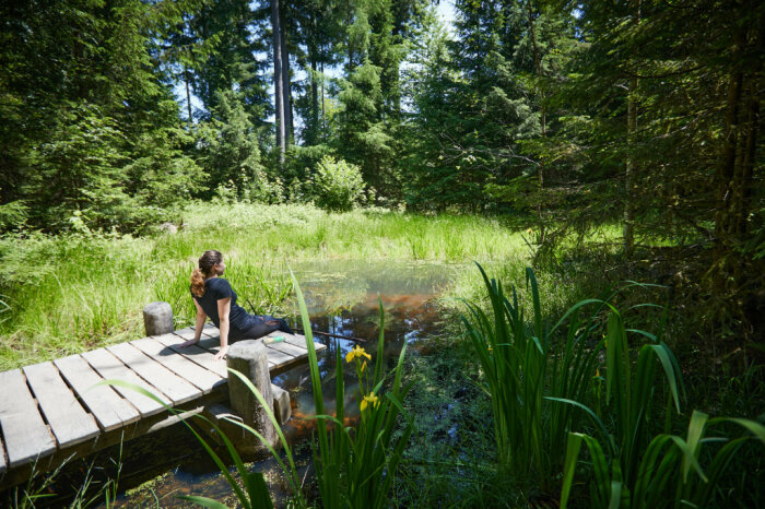 Frau sitzt auf einem Holzsteg an einem kleinen See im Wald.