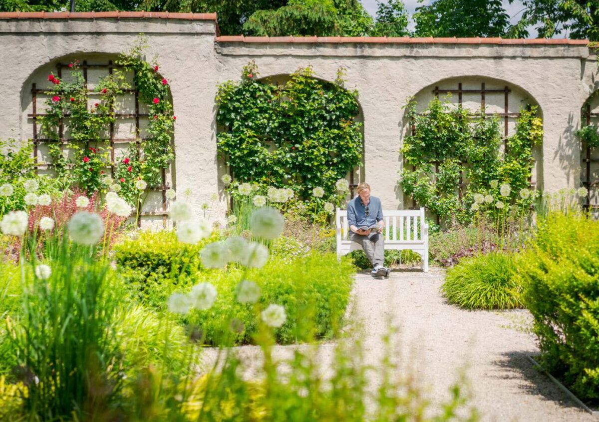 Mann sitzt in einem Schlossgarten auf einer weißen Bank und liest ein Buch.