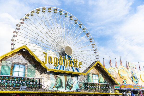 Volksfeststimmung. Im Vordergrund steht eine Jagdhütte im Hintergrund ein Riesenrad mit vielen Gondeln.