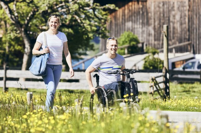 Zwei Personen, eine davon im Rollstuhl, auf einem Weg vor einer Wiese