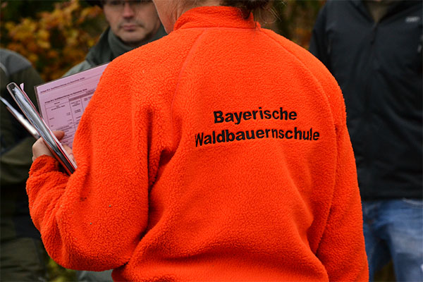 Rückansicht einer Person mit ornger Jacke. Aufschrift: Bayerische Waldbauernschule.