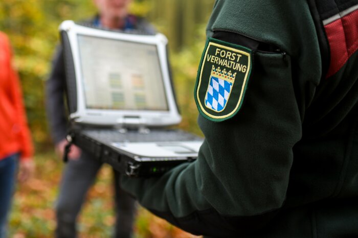 Person in Dienstkleidung der Bayerischen Forstverwaltung mit Wappen auf dem Arm hält Laptop in der Hand