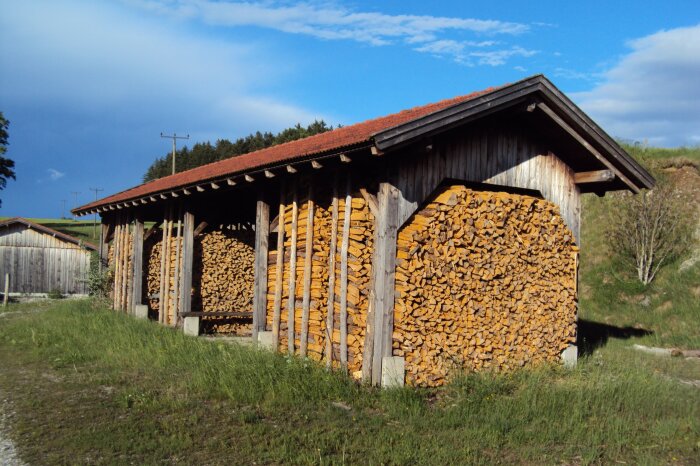 In einer an den Seiten offenen Hütte auf einer Wiese ist sauber bis unters Dach Brennholz aufgestapelt