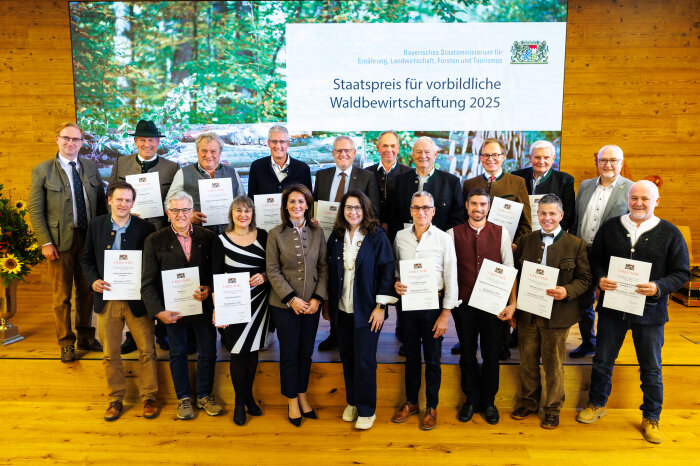 Gruppenfoto: Die Preisträgerinnen und Preisträger mit ihren Urkunden, im Hintergrund eine große Leinwand mit Waldbild und Schriftzug "Staatspreis für vorbildliche Waldbewirtschaftung 2025"