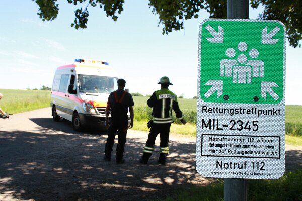 Nahaufnahme des Schildes an einem Rettungstreffpunkt; im Hintergrund zwei Personen und ein Krankenwagen (Foto: Martin Kolbe).